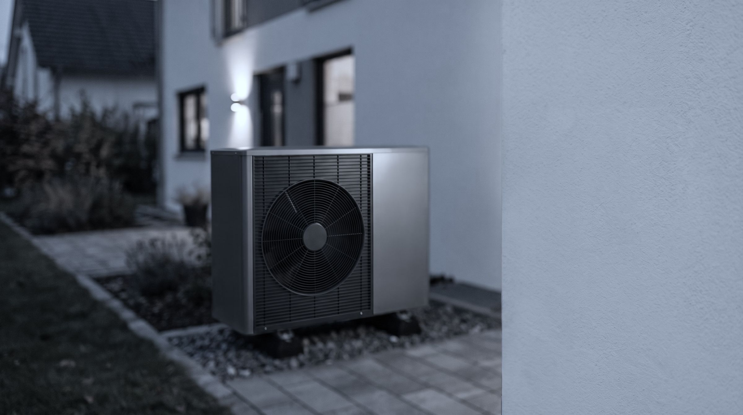 Außeneinheit einer Luft-Wärmepumpe auf Kiesbett vor einem hellen Wohnhaus in der Abenddämmerung