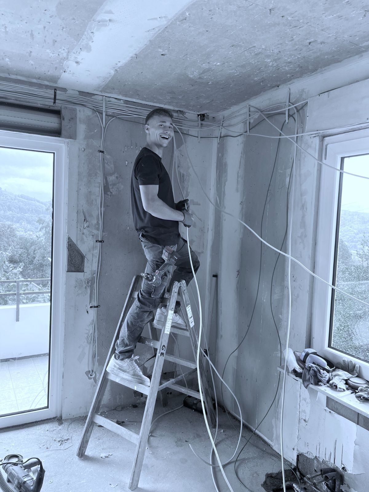 Ein Elektriker von Gerhards steht auf einer Leiter und verlegt Leitungen in einem Rohbauraum mit großen Fenstern.
